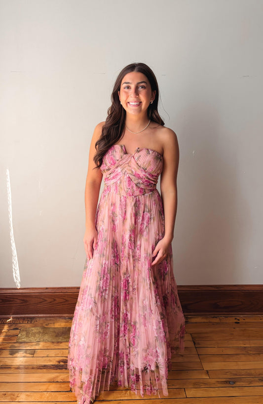 Woman wearing a pink floral dress standing against a plain wall.