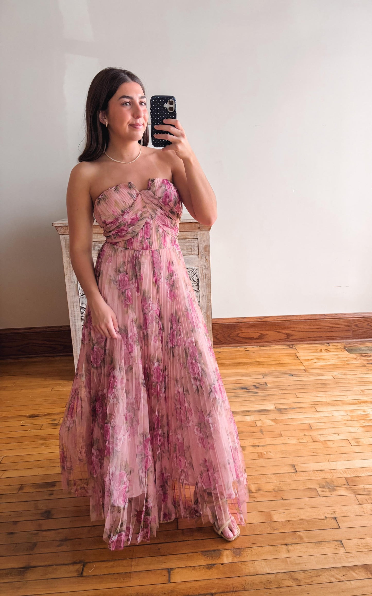 Woman in a floral dress taking a mirror selfie in a room with wooden flooring.