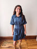 A woman wearing a short-sleeve button-front shirt dress in a medium blue denim-like fabric, paired with tan knee-high boots, standing on a wooden floor against a plain off-white wall.