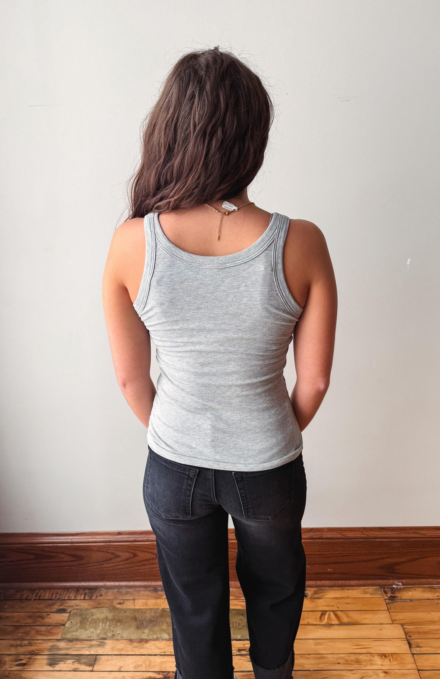 A woman viewed from behind wearing a light gray ribbed tank top and dark jeans, standing on a wooden floor against a plain off-white wall.
