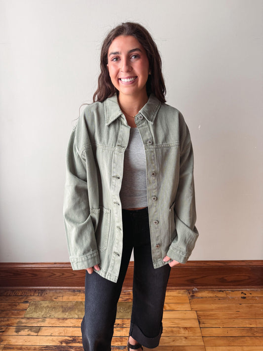 A woman smiling while wearing an oversized sage green denim jacket over a gray ribbed top and black jeans, standing in front of a plain off-white wall on a wooden floor.