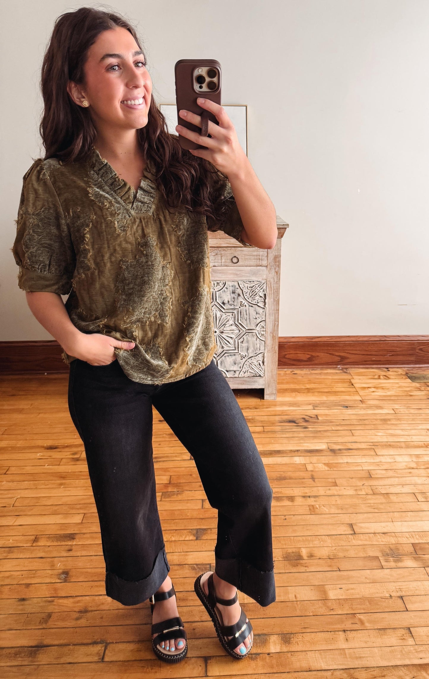 A woman takes a mirror selfie in a room with hardwood floors, wearing a short-sleeved olive green and gold patterned blouse, black wide-leg cropped pants, and black sandals.