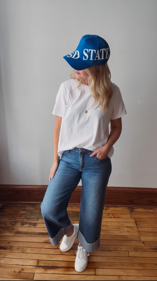 A woman wearing a blue baseball cap with 'USA STATE' printed on it, a white t-shirt, blue jeans, and white sneakers, standing on a wooden floor against a plain wall.