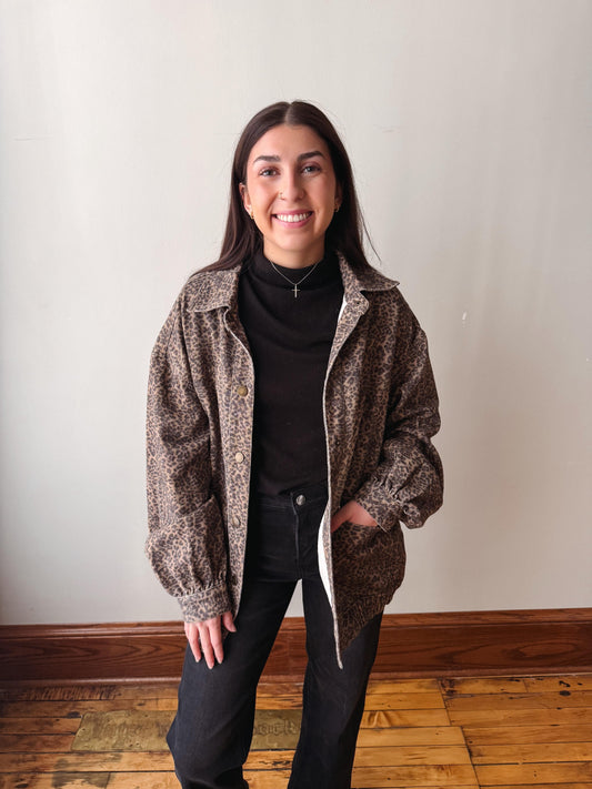 A woman modeling a brown and black patterned zip-up jacket over a black turtleneck and black pants, standing against a plain off-white wall with a wooden floor.