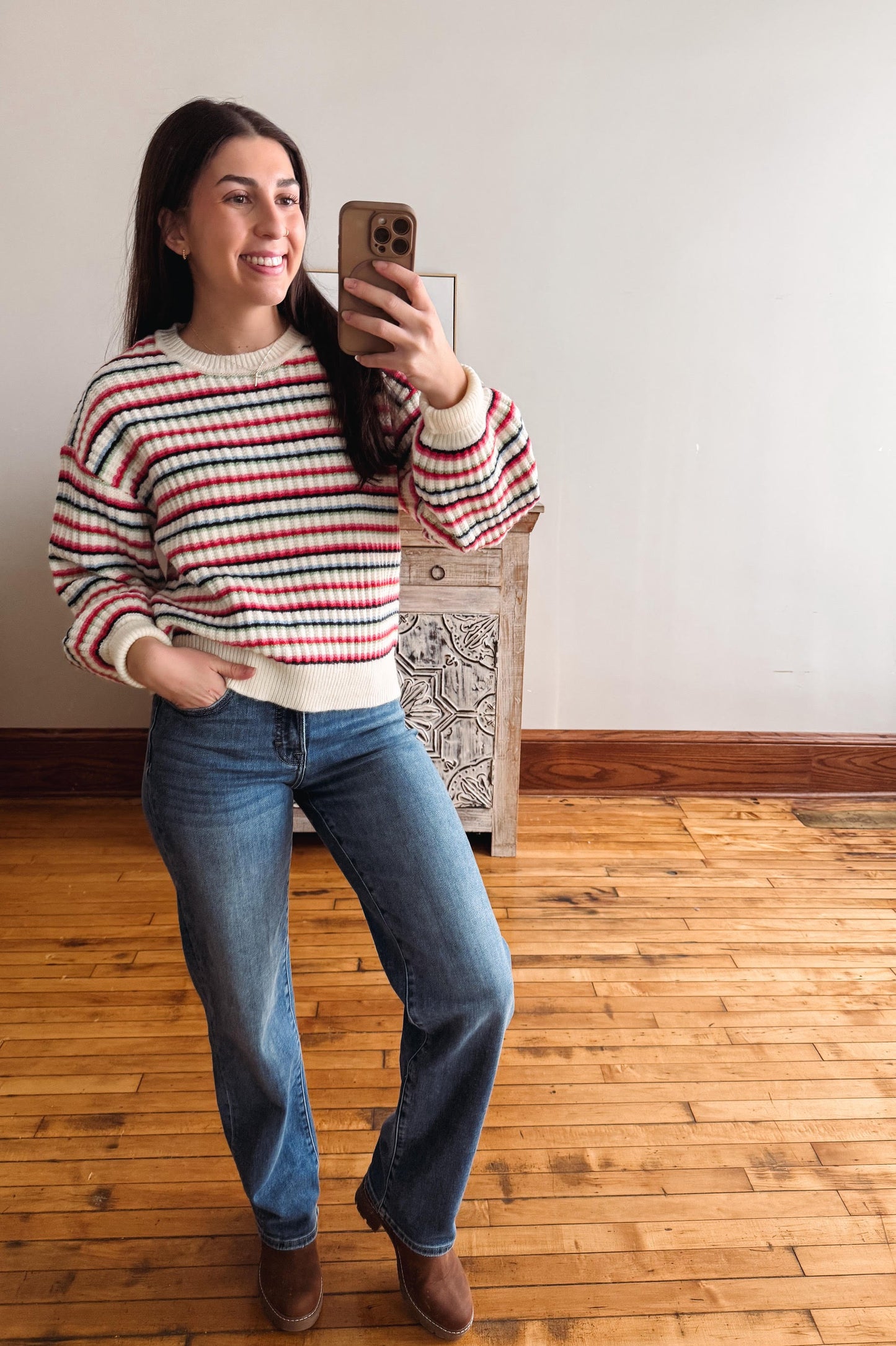 A woman taking a mirror selfie in a room with a light-colored wall and wooden floor, wearing a red, white, and black striped long-sleeve knit sweater, blue jeans, and brown boots.