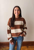 A woman wearing a brown and cream striped knit sweater with blue jeans, standing against a plain off-white wall with wood trim and a wooden floor.