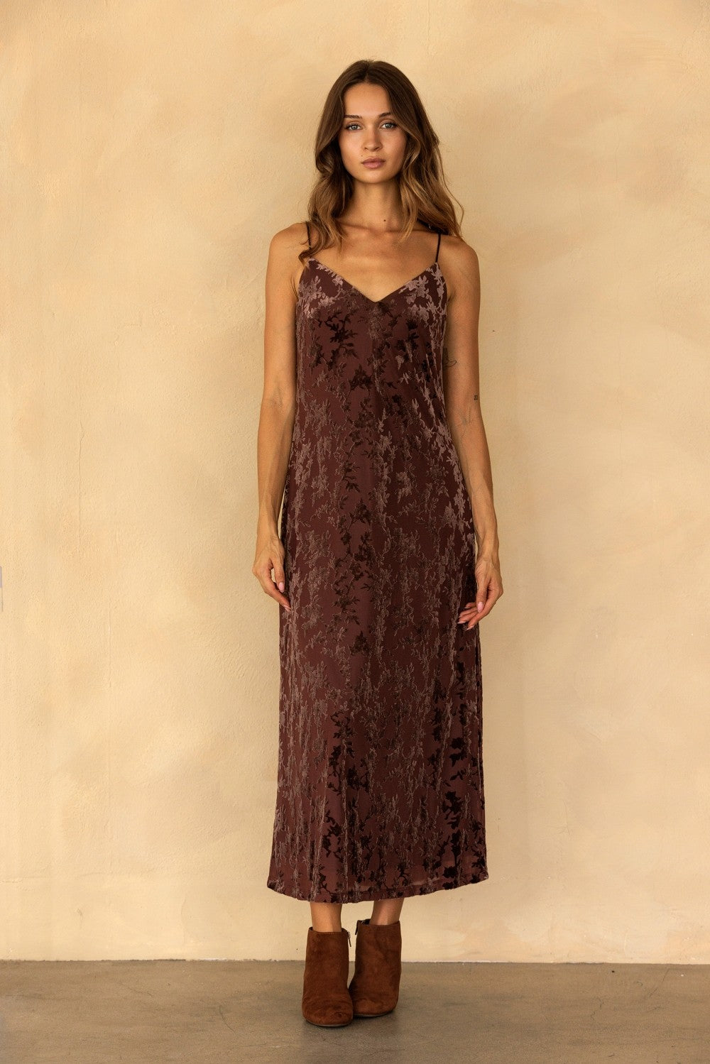 A woman modeling a dark brown velvet slip dress with a tonal floral jacquard pattern, paired with brown suede ankle boots, standing against a textured beige wall.