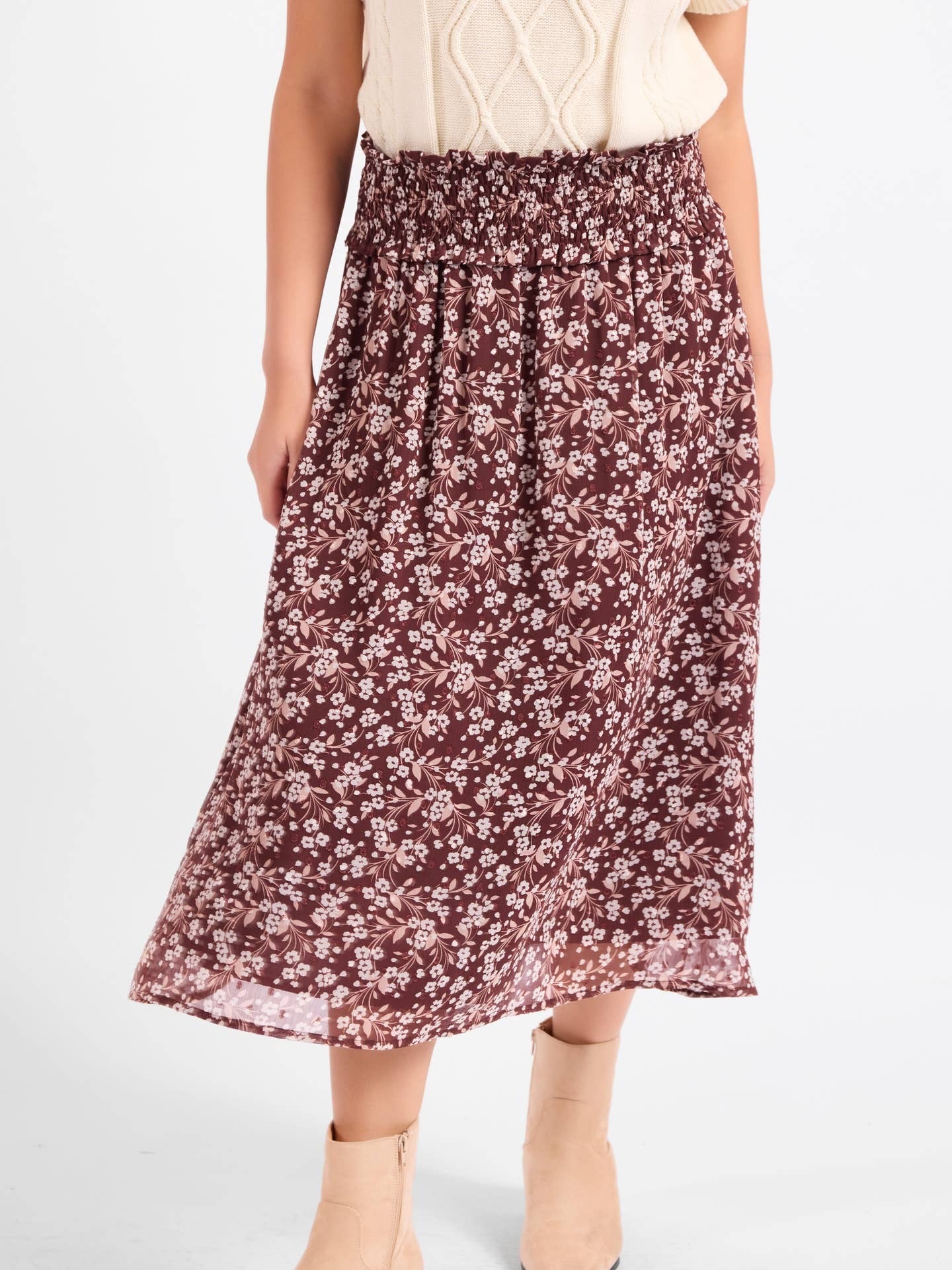 A woman wearing a mid-length, flowy skirt with a dense small floral print in dark brown and white. The skirt has an elastic waistband and is styled with a cream-colored knit top and beige ankle boots against a plain white studio background.