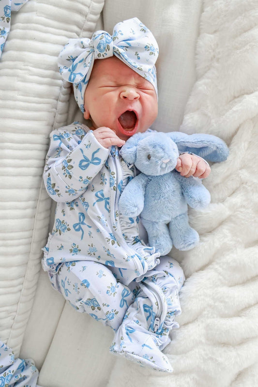 A newborn baby wearing a light blue and white patterned footed sleeper and matching bow headband, holding a light blue plush bunny toy while lying on a soft cream blanket.