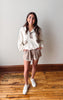 Woman wearing a white blouse and beige shorts standing on a wooden floor.