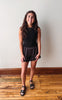 A young woman with long brown hair stands in a room with a light gray wall and wooden floor, wearing a black sleeveless top, black lace-trimmed shorts, and black sandals.