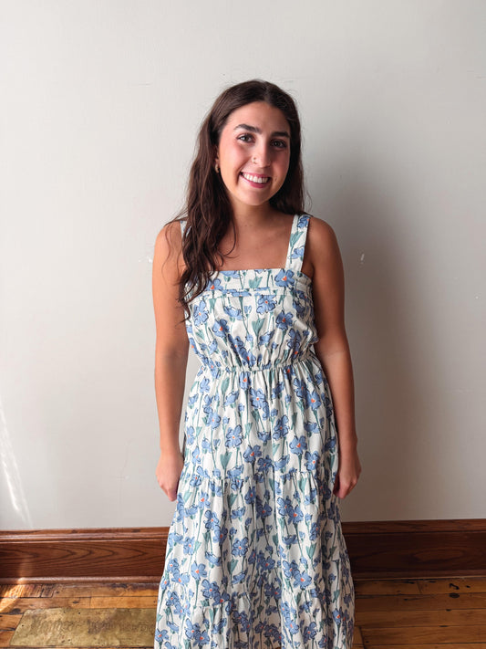 A young woman smiling and wearing a sleeveless, square-neck midi dress with a blue floral pattern on a cream background, standing against a plain off-white wall with wooden trim and a hardwood floor.