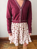 A person wearing a burgundy cardigan with white contrast stitching over a cream tiered skirt with a pink floral print, paired with tan boots, standing on a wooden floor against a plain wall.
