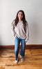 A woman stands in a room with a light gray wall and wooden floor, wearing a light beige long-sleeve crewneck sweatshirt, blue jeans, and white slide sandals.