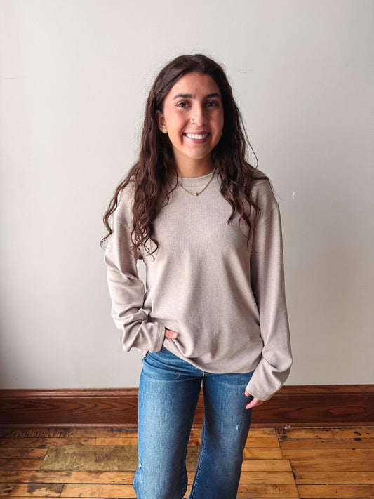 A smiling young woman with long wavy brown hair stands indoors wearing a light taupe-beige long-sleeve knit sweater and blue jeans against a plain off-white wall with a wooden floor.