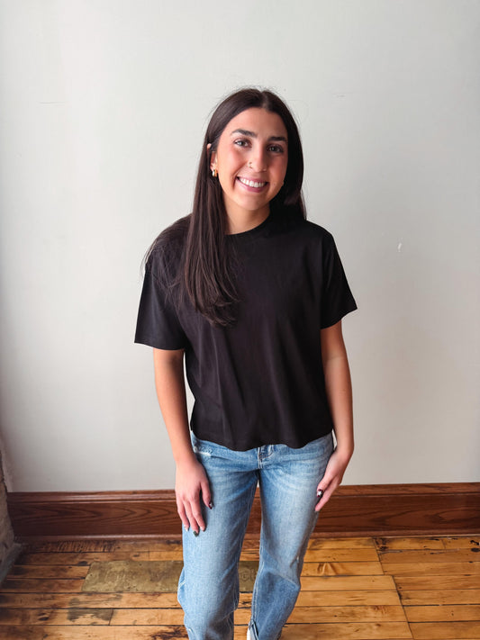 A woman with long dark hair stands indoors wearing a black short-sleeve crewneck t-shirt and light wash blue jeans, posing against a plain off-white wall with a wooden floor visible.