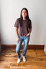 A woman stands in a room with a light gray wall and wooden floor, wearing a dark brown short-sleeve t-shirt, blue jeans, and white sneakers.