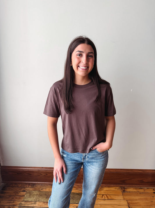 A woman with long dark hair stands indoors wearing a short-sleeve crewneck t-shirt in a muted brownish-purple color and light-wash blue jeans, posing against a plain off-white wall with wooden flooring.