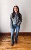 A woman stands on a wooden floor against a plain off-white wall, wearing a gray knit button-front cardigan over a denim shirt, blue jeans, and brown boots.