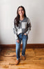 A woman wearing a gray and white checkered V-neck sweater, blue jeans, and brown suede boots, standing on a wooden floor against a plain off-white wall.