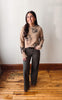 A woman stands in a room with a white wall and wooden floor, wearing a beige knit sweater with black floral embroidery, dark gray wide-leg pants, and a black belt.