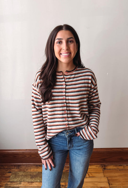 A woman with long dark hair stands indoors wearing a cropped, long-sleeve knit top with horizontal stripes in brown, beige, and white, paired with blue jeans.