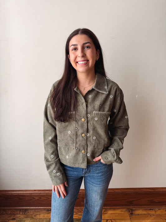 A woman with long dark hair stands indoors wearing a muted olive green corduroy button-up shirt with white embroidered details and blue jeans, set against a plain off-white wall and wooden floor.