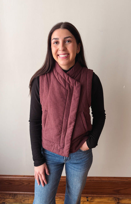 A woman wearing a dusty rose wrap-style quilted vest over a black long-sleeve shirt and blue jeans, standing against a plain off-white wall.