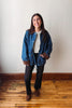 A woman stands in a room with a beige wall and wooden floor, wearing a blue denim jacket with brown suede-like trim over a cream top and black pants.