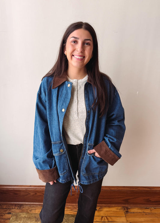 A woman wearing a blue denim jacket with brown corduroy collar and cuffs, layered over a light gray henley top and black pants, standing against a plain beige wall.