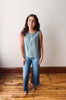 Woman wearing a light blue sleeveless top and jeans standing on a wooden floor.