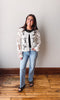 Woman wearing a white floral jacket and blue jeans standing on a wooden floor against a plain wall.