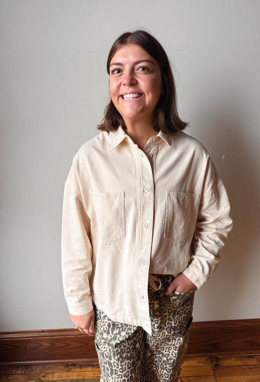 A woman smiling and looking up, wearing a light beige long-sleeve button-up shirt and leopard-print pants, standing against a plain light gray wall with a wooden baseboard and floor.