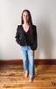 Woman wearing a black blouse and blue jeans standing on a wooden floor with a white wall background