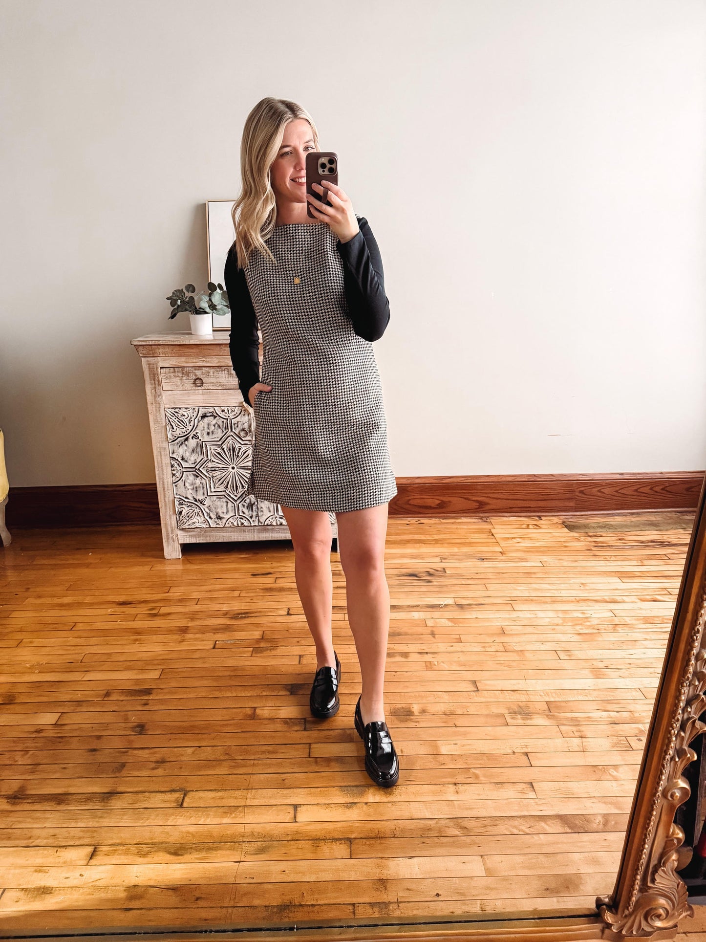 Woman taking a mirror selfie wearing a gray checkered dress and black shoes in a room with wooden flooring.
