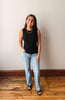 Woman wearing a black sleeveless top and light blue jeans standing on a wooden floor against a white wall.