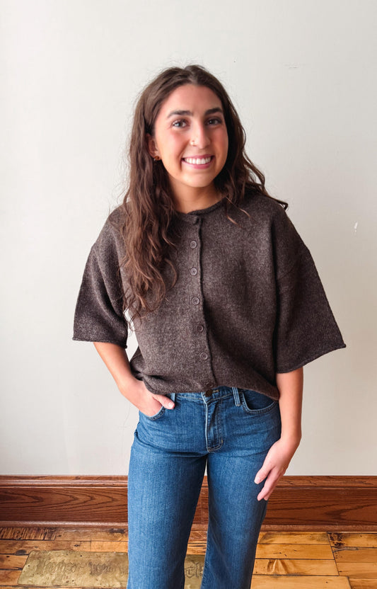 A woman with long brown hair smiling, wearing a dark brown short-sleeve button-up knit top and blue jeans, standing against a plain off-white wall with a wooden baseboard and floor.