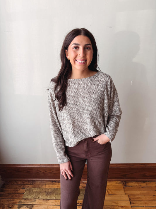 A woman with long dark hair stands indoors wearing a textured, heathered gray and white long-sleeve knit top and dark brown pants, posing against a plain off-white wall with wooden flooring.