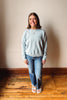 A woman stands on a wooden floor against a beige wall, wearing a light blue crewneck sweatshirt, blue jeans, and white sandals.