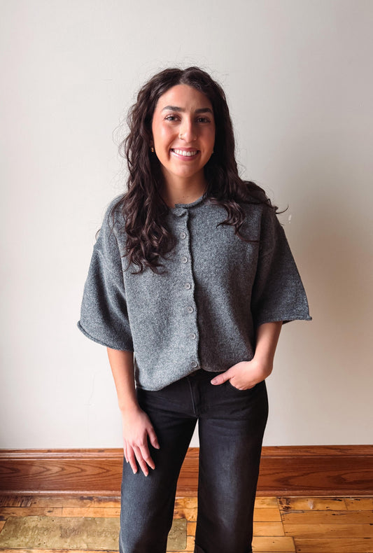 A woman with long dark curly hair stands indoors wearing a gray short-sleeve button-front cardigan or shrug and dark jeans, with one hand in her pocket. The background is a plain off-white wall with a wooden baseboard and wood floor.
