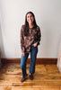 A woman stands in a room with a white wall and wooden floor, wearing a brown long-sleeve button-up shirt with a white floral print, blue jeans, and brown loafers.