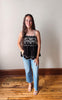 Woman wearing a black sleeveless top with white lace details and blue jeans standing on a wooden floor.