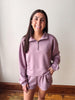 A woman modeling a matching dusty purple lounge set consisting of a quarter-zip sweatshirt and matching shorts, standing against a plain off-white wall with wooden flooring visible at the bottom.