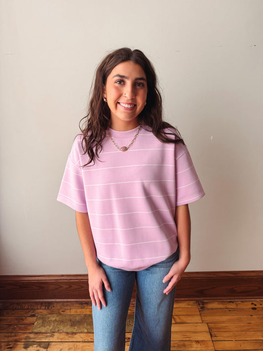 Person wearing a pink striped shirt and blue jeans standing against a plain wall.