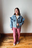 A woman stands on a wooden floor against a plain wall, wearing a light blue denim jacket with ruffle details on the shoulders and sleeves, paired with dusty rose pants and white sneakers.