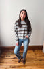 A woman stands in a room with a white wall and wooden floor, wearing a gray and brown horizontally striped knit sweater, blue jeans, and brown loafers.