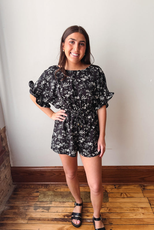Woman wearing a black floral romper standing in a room with wooden flooring and a white wall.