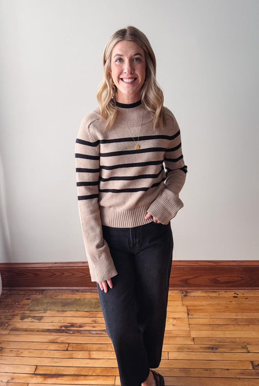 Woman wearing a beige sweater with black stripes and dark pants indoors.