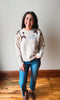 A woman wearing a cream knit sweater with brown leaf details and a light blue flower on the chest, paired with blue jeans and brown slip-on shoes, standing in a room with a light gray wall and wooden floor.