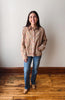 A woman stands in a room with a light gray wall and wooden floor, wearing a beige button-up shirt with a subtle floral pattern, blue jeans, and tan boots.